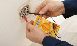 Elektryk sprawdza napięcie w gniazdku za pomocą multimetru. Na wyświetlaczu widnieje wartość 221V.