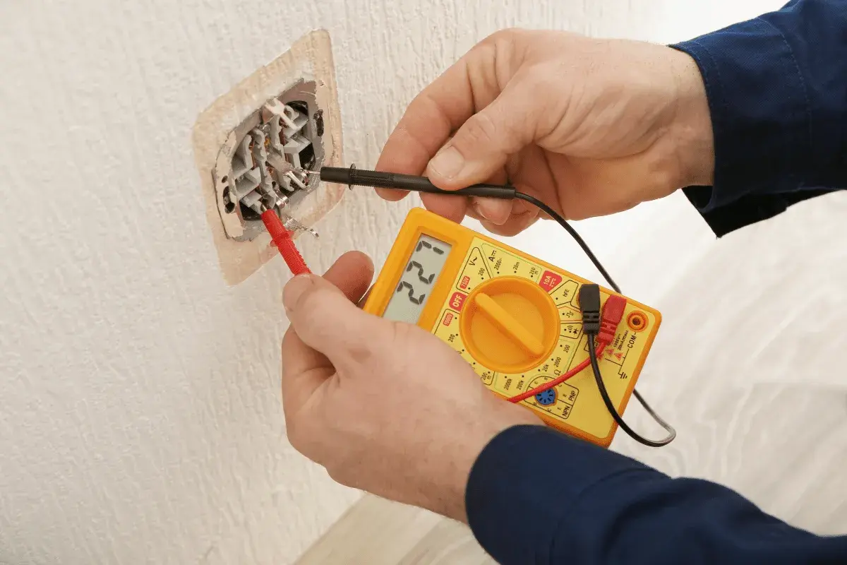 Elektryk sprawdza napięcie w gniazdku za pomocą multimetru. Na wyświetlaczu widnieje wartość 221V.